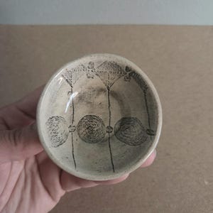 May include: A small, off-white ceramic bowl with a hand-drawn black floral design. The bowl's interior features three stylized flowers and spiral accents. The bowl is held in a hand, showcasing its size and design.