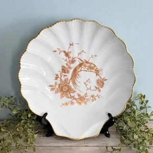May include: A white ceramic serving dish shaped like a seashell with a gold rim. The dish features a brown and gold floral design with a bird in the center.