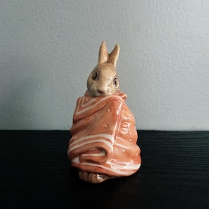 May include: A ceramic figurine of a brown rabbit wrapped in a peach-colored blanket with white stripes and small white dots. The rabbit has upright ears, dark eyes, and a neutral expression, with the blanket draped around its body in folds.