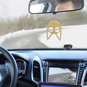 Op de afbeelding: Een gouden glitter auto-hanger in de vorm van een gestileerde uil, hangend aan een achteruitkijkspiegel. De hanger heeft uitgesneden ogen en een lachende mond. Het dashboard van de auto en een besneeuwde weg zijn zichtbaar op de achtergrond.