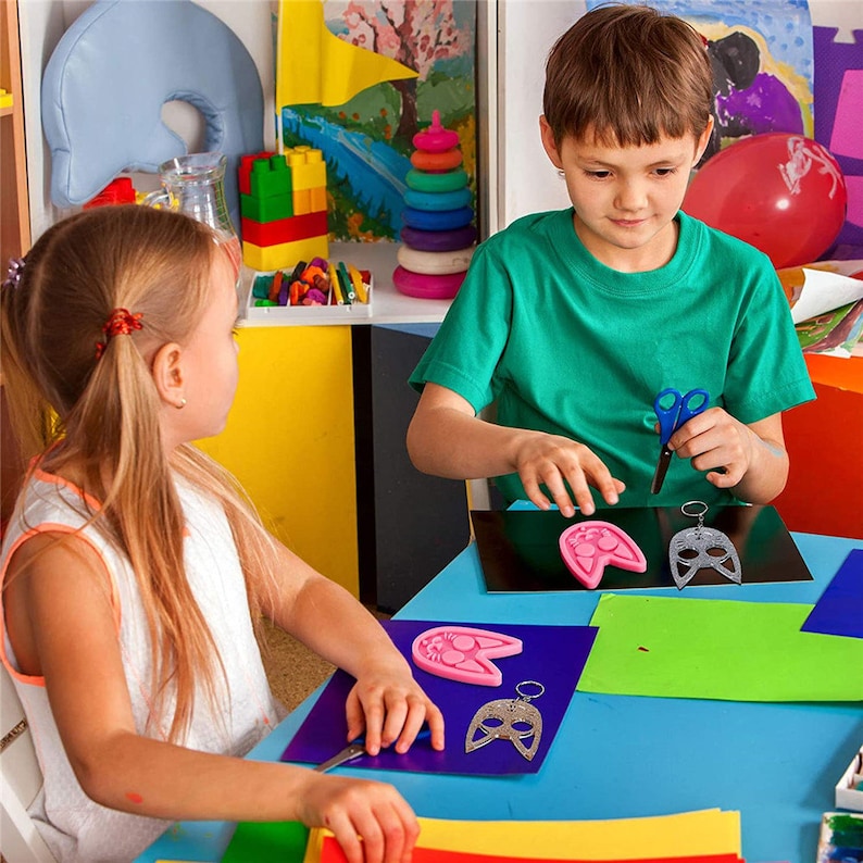 Op de afbeelding: Twee kinderen zitten aan een blauwe tafel en zijn aan het knutselen. E&eacute;n kind houdt een schaar vast, de ander werkt met roze en zilveren maskervormen. Op de achtergrond kleurrijk papier en speelgoed, wat wijst op een creatieve omgeving.