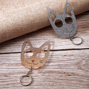 Op de afbeelding: Twee katvormige sleutelhangers met glinsterende oppervlakken. E&eacute;n is zilver, de andere is goud. Elke sleutelhanger heeft een ronde ring. De sleutelhangers liggen op een houten oppervlak.