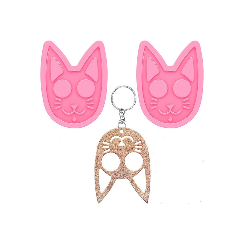 Op de afbeelding: Een roze siliconen katvormige mal en een gouden glitter katten sleutelhanger met een zilveren sleutelring. De mal heeft een kattengezicht ontwerp met grote ronde ogen, snorharen en een kleine neus. De sleutelhanger heeft een vergelijkbaar kattengezicht ontwerp.