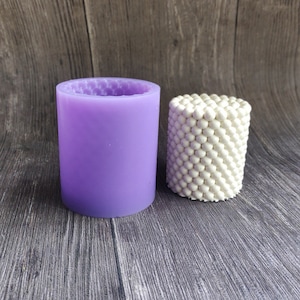 May include: A cylindrical, lavender-colored silicone mold with a honeycomb pattern on the rim, next to a white, textured candle. The candle has a spherical, bubble-like design. The items are on a gray wooden surface.
