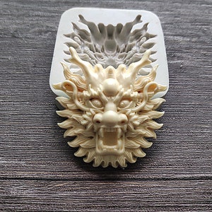 Puede incluir: Un molde de silicona beige con forma de cabeza de dragón con detalles intrincados. El dragón tiene la boca abierta, dientes afilados y rasgos faciales detallados. El molde está en un marco rectangular blanco.