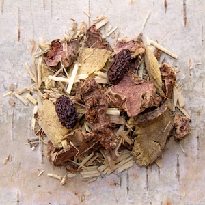 May include: A close-up of a mixture of dried herbs and roots, including pieces of liquorice root, angelica root, and dried berries. The mixture is arranged on a light brown surface.