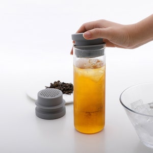 Peut inclure: Un infuseur à thé en silicone gris avec un filtre en maille se trouve sur une assiette blanche à côté d'une bouteille en verre transparente remplie de thé glacé. La bouteille a un couvercle en silicone gris.