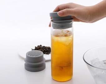 May include: A grey silicone tea infuser with a mesh filter sits on a white plate next to a clear glass bottle filled with iced tea. The bottle has a grey silicone lid.