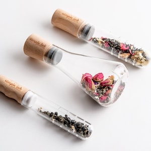 Puede incluir: Tres infusores de té de vidrio con tapas de madera. Los infusores están llenos de hojas de té secas y flores. Los infusores están etiquetados como "simple lab experience".