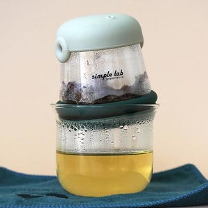 May include: A clear glass tea infuser with a green silicone lid and a green silicone ring. The infuser is filled with loose leaf tea and is sitting on top of a clear glass cup filled with brewed tea. The cup is sitting on a blue towel.