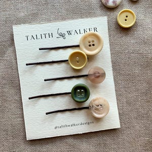 May include: Five bobby pins with attached buttons in various colours and textures, including cream, yellow, translucent pink, dark green, and marbled beige, displayed on a white card labelled TALITH WALKER and @talithwalkerdesigns.