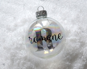 Peut inclure: Boule de Noël en verre transparent avec un capuchon argenté. L'ornement porte le nom "romane" et la lettre "R" en script noir. La boule irisée repose sur une surface blanche et enneigée.