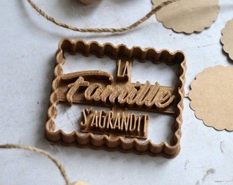 May include: A brown plastic cookie cutter shaped like a rectangle with the words "La Famille S'Agrandit" in a decorative font.