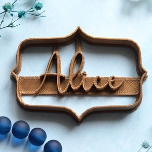 May include: A brown plastic cookie cutter in a decorative frame shape, with the name "Alice" in a cursive font. The cutter is on a light blue surface, with blue decorative elements.