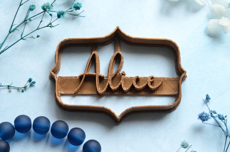 May include: A brown plastic cookie cutter in a decorative frame shape, with the name "Alice" in a cursive font. The cutter is on a light blue surface, with blue decorative elements.