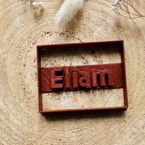 Könnte beinhalten: Ein brauner Plastik-Ausstecher in Form eines Rechtecks mit dem Namen "Elliam" in erhabenen Buchstaben.