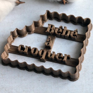 May include: A brown plastic cookie cutter with a wavy edge. The cutter is shaped like a rectangle and has the words "Bebe a croquer" embossed on it.