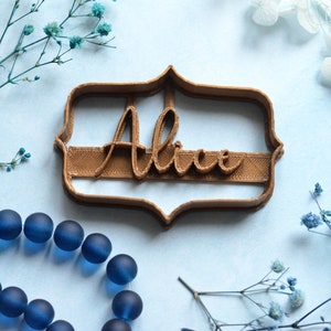 May include: A brown cookie cutter in the shape of a decorative frame with the name "Alice" in cursive script. The cutter is on a light blue surface, with blue beads and sprigs of blue flowers.