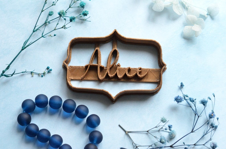 May include: A brown cookie cutter in the shape of a decorative frame with the name "Alice" in cursive script. The cutter is on a light blue surface, with blue beads and sprigs of blue flowers.