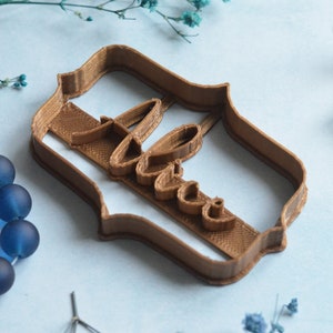 May include: A brown cookie cutter in the shape of a decorative frame with the word "Love" in cursive script. The cutter is made of plastic and sits on a light blue surface, with blue beads and flowers in the background.