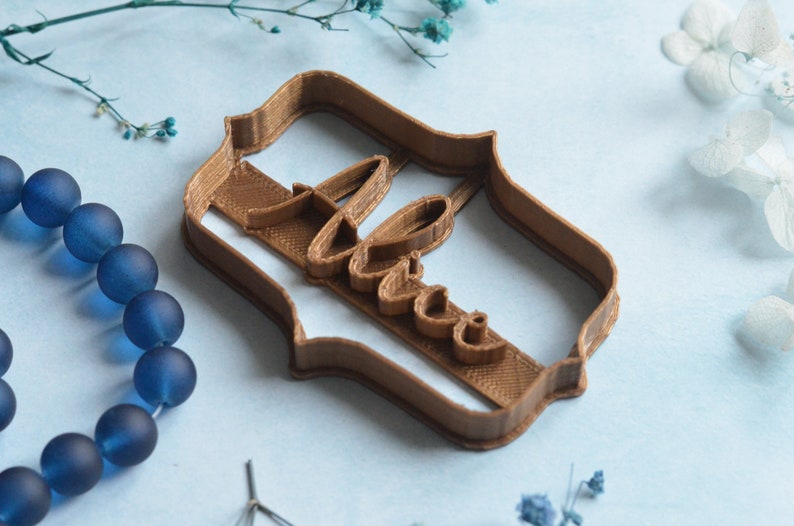 May include: A brown cookie cutter in the shape of a decorative frame with the word "Love" in cursive script. The cutter is made of plastic and sits on a light blue surface, with blue beads and flowers in the background.