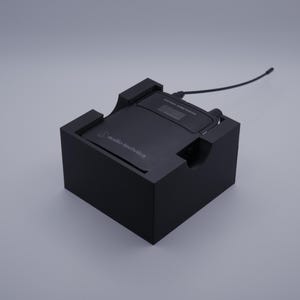 May include: A black audio-technica wireless microphone receiver is placed in a custom black holder. The receiver has a small display screen and a black cable extending from the top. The holder has cutouts for the receiver and cable.