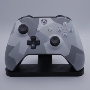May include: An Xbox controller with a grey camouflage design, on a black stand. The controller has the Xbox logo, buttons, and a directional pad. The stand is a simple, rectangular shape.