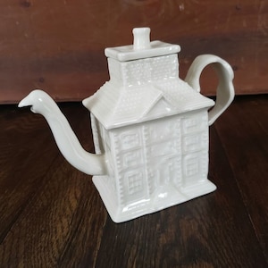 Peut inclure: Théière en céramique blanche en forme de maison, avec cheminée, toit et façade détaillée avec fenêtres et porte. Le bec verseur est incurvé et la poignée est une boucle. La théière est posée sur une surface en bois.