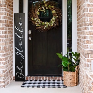 May include: A black wooden sign with white calligraphy that reads "Oh, hello" leaning against a brick wall next to a black front door with a green and brown wreath. A potted plant sits on the porch in a woven basket.