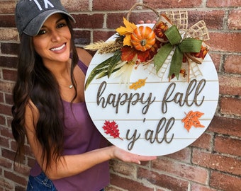 May include: A white round wooden sign with the text "happy fall y'all" in brown lettering. The sign is decorated with fall-themed elements, including a small orange pumpkin, fall leaves, and a burlap bow.