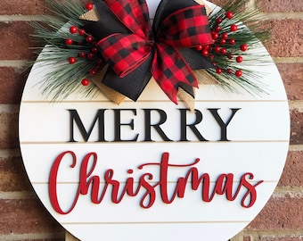 Może przedstawiać: Okrągły drewniany znak w kolorze białym z napisem "Merry Christmas" w czarnych i czerwonych literach. Znak jest ozdobiony czarną i czerwoną kokardą w kratę, zielonymi gałązkami sosny i czerwonymi jagodami. Znak jest zawieszony na sznurku i opiera się o drewniany sztalug.