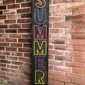 May include: A dark brown wooden sign with the word "SUMMER" in large, colorful letters. The letters are outlined in various colors, including orange, blue, pink, yellow, and purple. The words "Summer Vibes" are written in teal at the bottom.