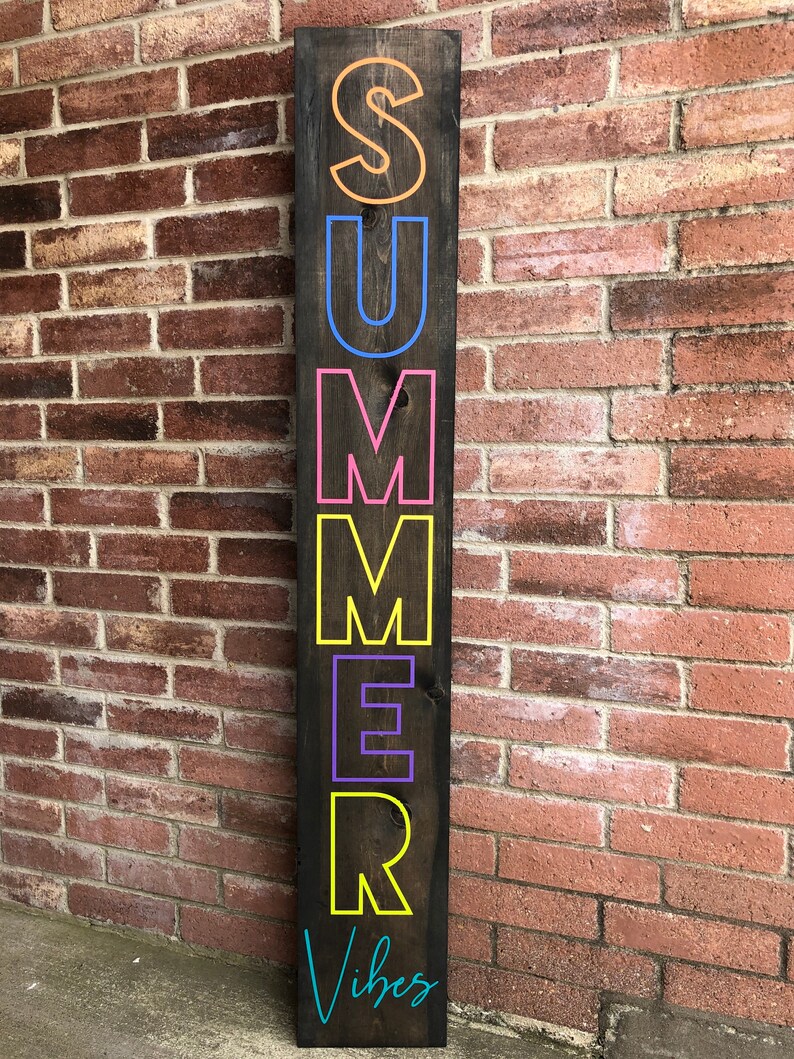 May include: A dark brown wooden sign with the word "SUMMER" in large, colorful letters. The letters are outlined in various colors, including orange, blue, pink, yellow, and purple. The words "Summer Vibes" are written in teal at the bottom.