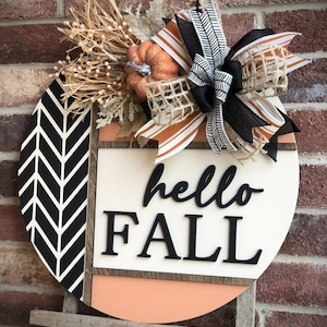 May include: A round wooden sign with a black and white chevron pattern, a peach colored background, and the words "hello fall" in black lettering. The sign is decorated with a burlap bow, a small orange pumpkin, and fall-themed greenery.