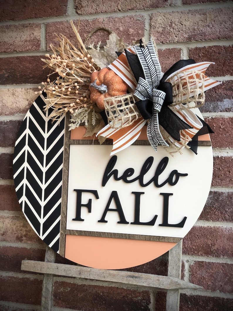 May include: A round wooden sign with a black and white chevron pattern, a peach colored background, and the words "hello fall" in black lettering. The sign is decorated with a burlap bow, a small orange pumpkin, and fall-themed greenery.