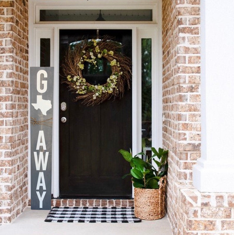May include: A gray wooden sign with the text "G'AWAY" in white letters. The sign is decorated with a white outline of the state of Texas. The sign is hanging on a brick wall next to a black door with a wreath.