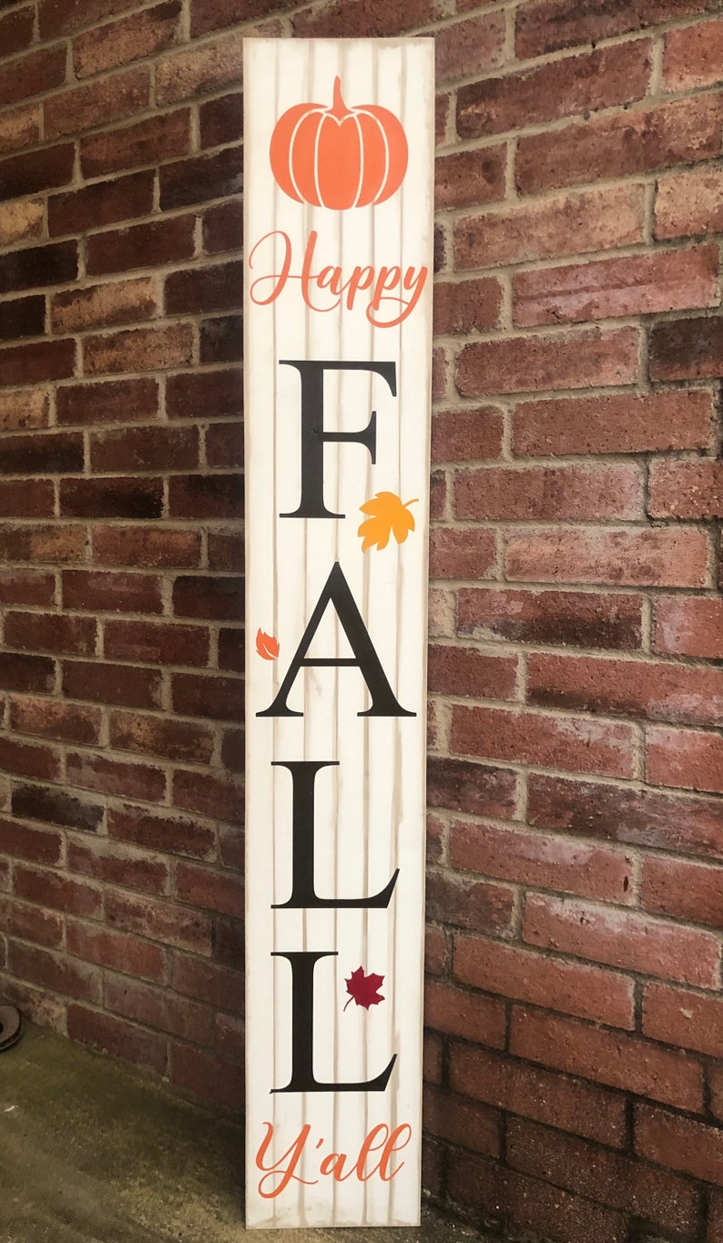 May include: A white wooden sign with the words "Happy Fall Y'all" in orange and black lettering. The sign features a pumpkin and fall leaves.