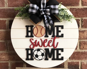 以下が含まれることがあります： 白地に黒と赤の文字で「Home Sweet Home」と書かれた丸い木製ドアハンガー。デザインには野球ボールとサッカーボールが組み込まれています。黒と白のチェック柄のリボンに緑の葉が付けられています。
