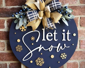 May include: A navy blue round wooden sign with the words "let it snow" in white script. The sign is decorated with gold snowflakes and dots, and a large bow with gold and plaid ribbons. The sign is hanging on a brick wall.