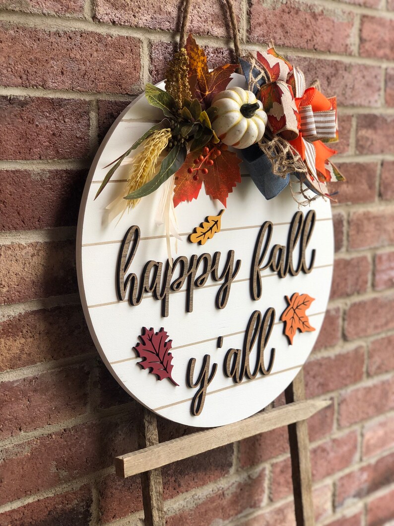 Puede incluir: Un letrero redondo de madera blanca con las palabras "happy fall y'all" en letras marrones. El letrero est&aacute; decorado con adornos de oto&ntilde;o, incluyendo una calabaza blanca, hojas naranjas y rojas y un lazo de arpillera.