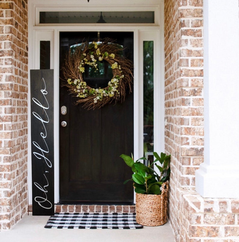 May include: A black wooden sign with white calligraphy that reads "Oh, hello" leaning against a brick wall next to a black front door with a green and brown wreath. A potted plant sits on the porch in a woven basket.
