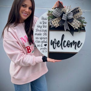May include: A round wooden welcome sign with a black and white design. The sign features the word "welcome" in black script, along with the text "This is the day the Lord has made: Let us rejoice & be glad in it. Psalm 118:24."