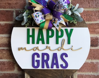 May include: A round wooden door hanger with a white background and a brown wood trim. The words "HAPPY mardi GRAS" are written in green, gold, and purple letters. A purple, gold, and green bow with white ribbon is attached to the top of the hanger.