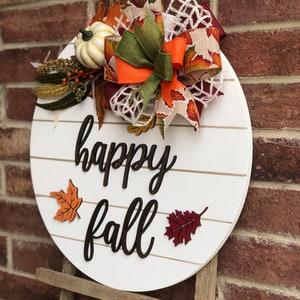 May include: White round wooden sign with the text "happy fall" in black script. The sign is decorated with a brown, orange, and green bow, a small white pumpkin, and fall-colored leaves.