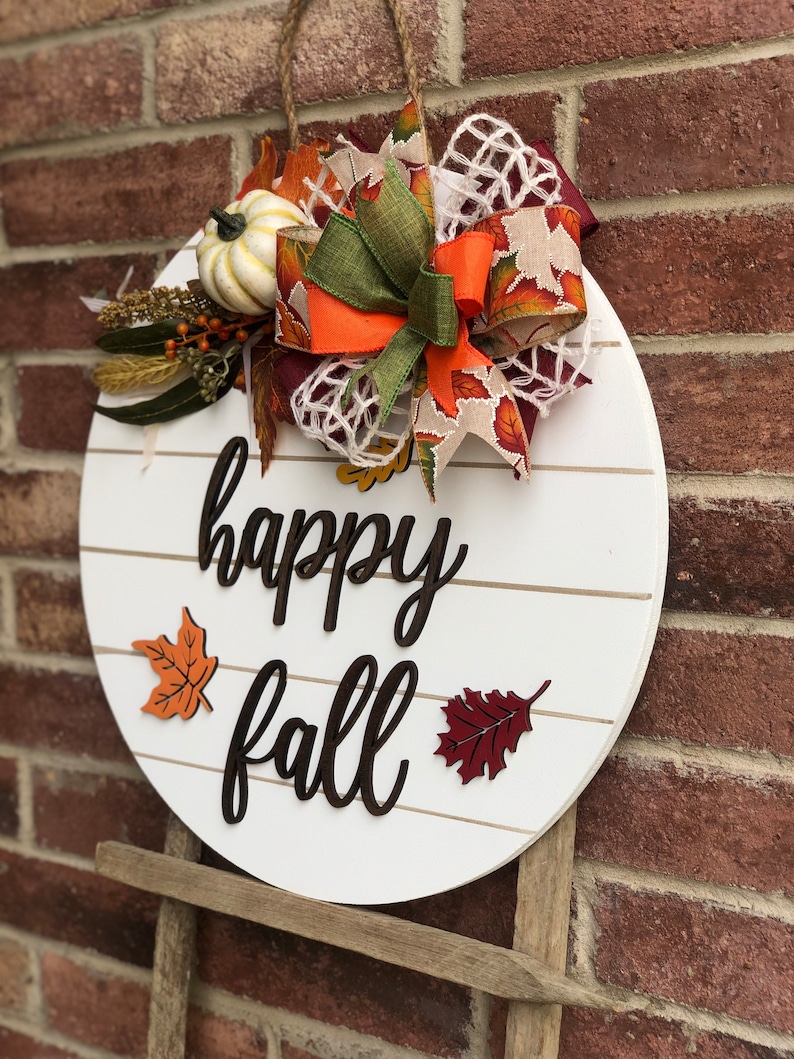 May include: White round wooden sign with the text "happy fall" in black script. The sign is decorated with a brown, orange, and green bow, a small white pumpkin, and fall-colored leaves.