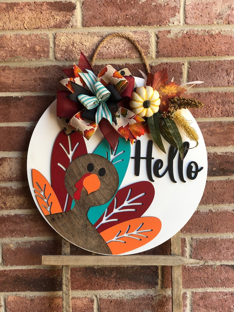 May include: A round wooden door hanger with a turkey design. The turkey is brown with a teal head and orange and red feathers. The door hanger also has a white bow with red, teal, and black ribbon. The door hanger has the word 'Hello' written on it in black lettering.