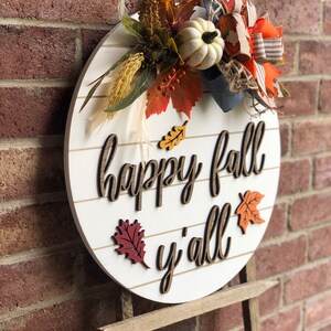 Puede incluir: Un letrero redondo de madera blanca con las palabras "happy fall y'all" en letras marrones. El letrero est&aacute; decorado con elementos de oto&ntilde;o, como una calabaza blanca, hojas naranjas y rojas y un lazo de arpillera.