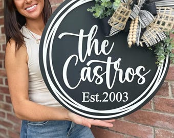 May include: A round black wooden sign with white lettering that reads "The Castros Est.2003". The sign is decorated with a bow, greenery, and a jute rope hanger. The sign is held by a person.