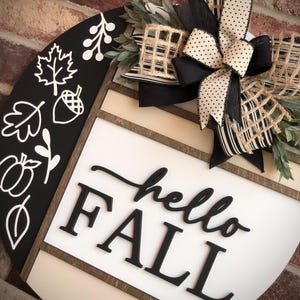 May include: A black and white round wooden sign with the words "hello fall" in black lettering. The sign is decorated with a black and white striped bow, a burlap bow, and a sprig of greenery.