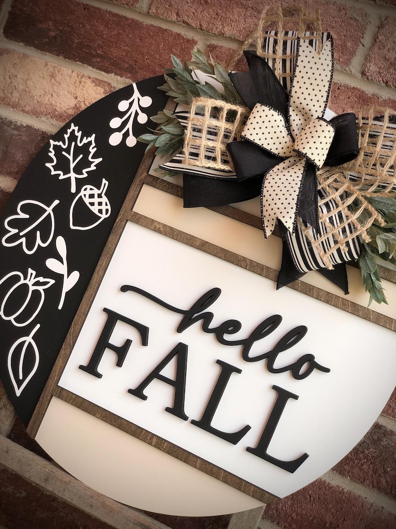 May include: A black and white round wooden sign with the words "hello fall" in black lettering. The sign is decorated with a black and white striped bow, a burlap bow, and a sprig of greenery.
